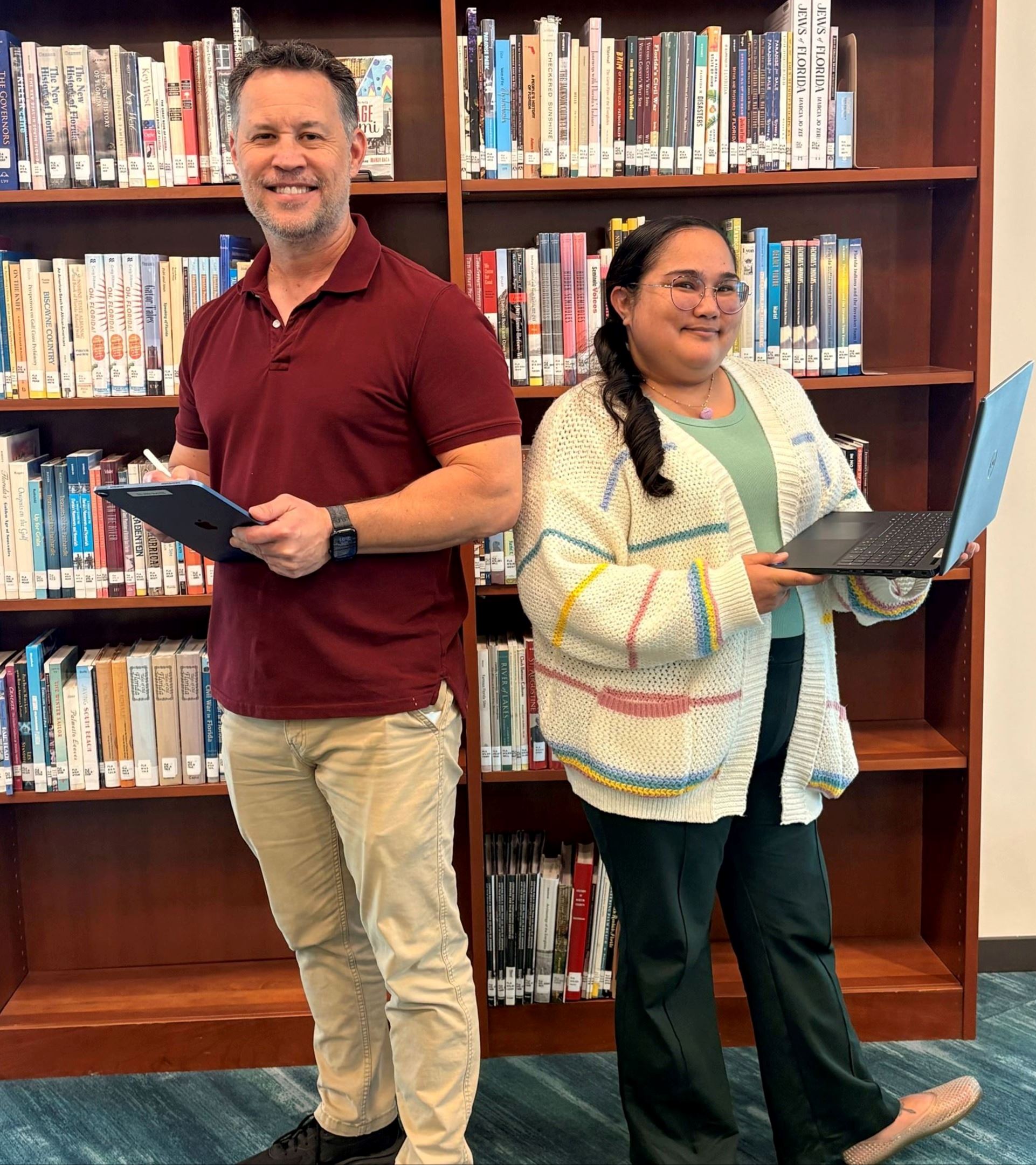 A male and female holding laptops, standing in front of book shelf