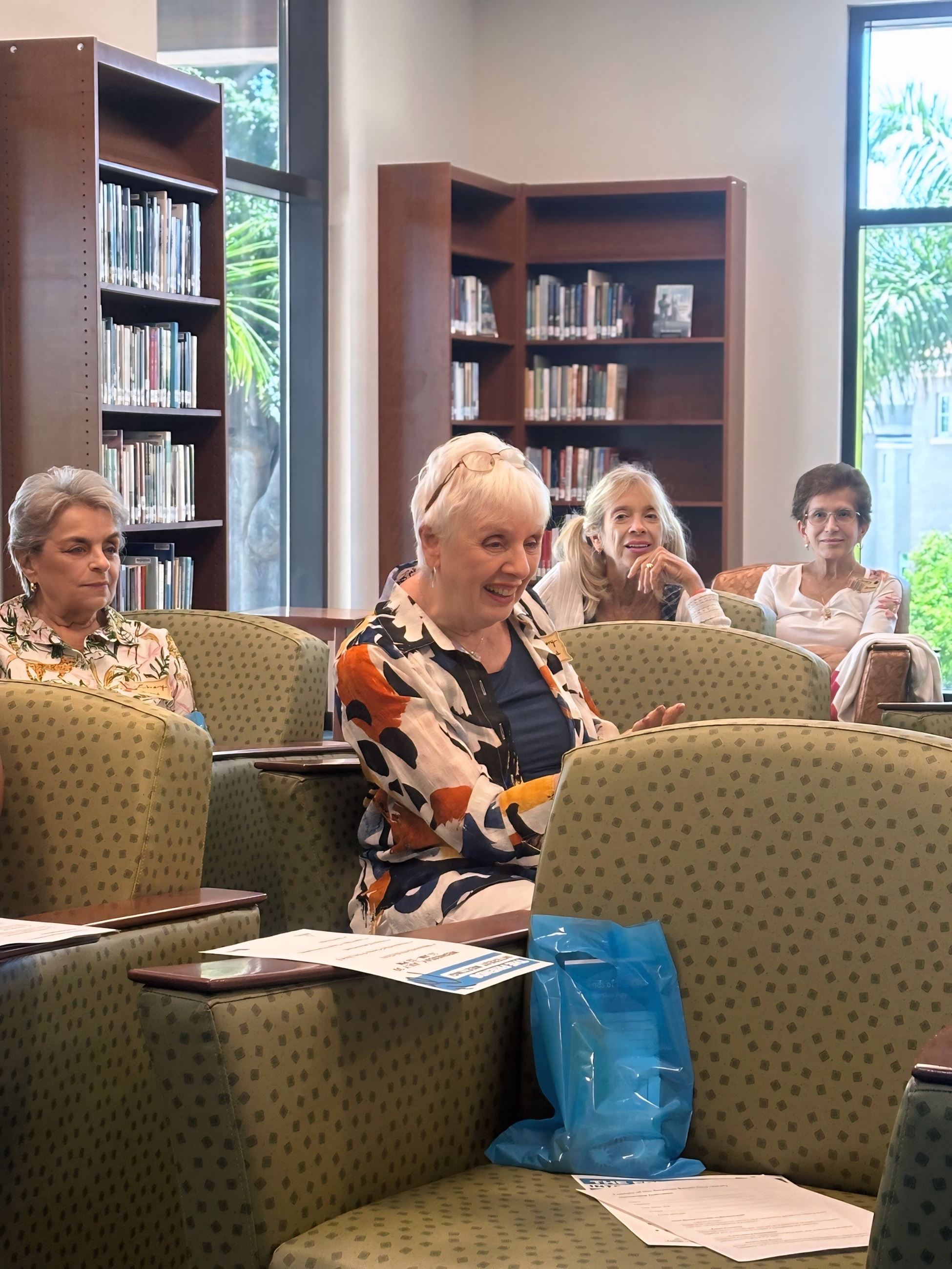 A group of people seated in a library meeting room with bookshelves and large windows showing greene